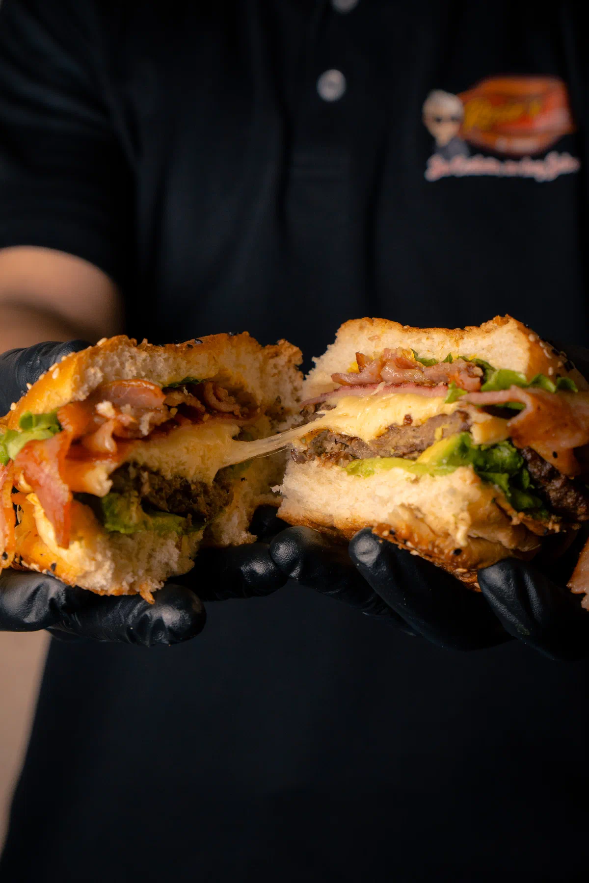 Hamburguesa partida con tocino y queso derretido, sostenida con guantes negros