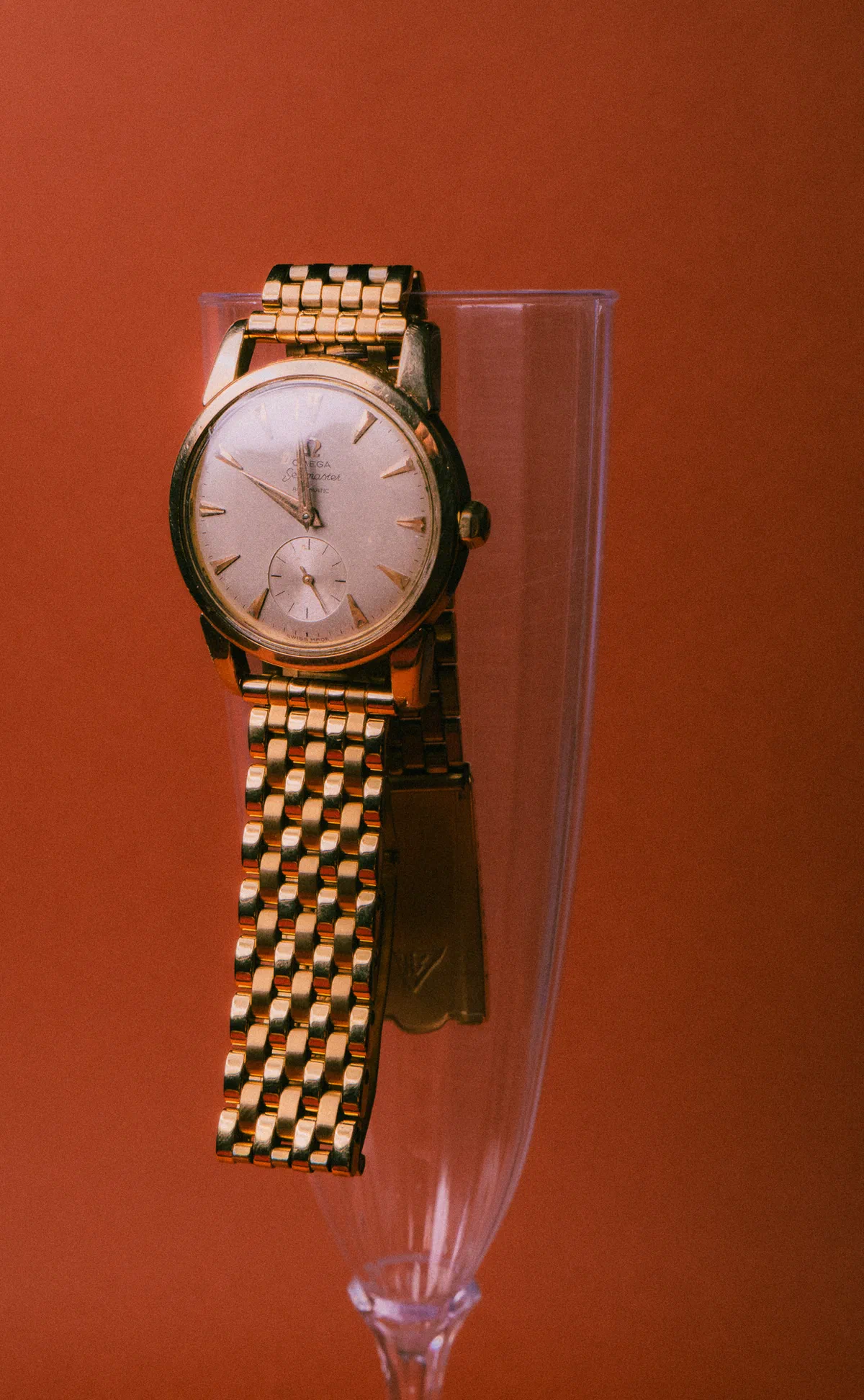 Reloj Omega dorado colgado de una copa de cristal, fondo rojo